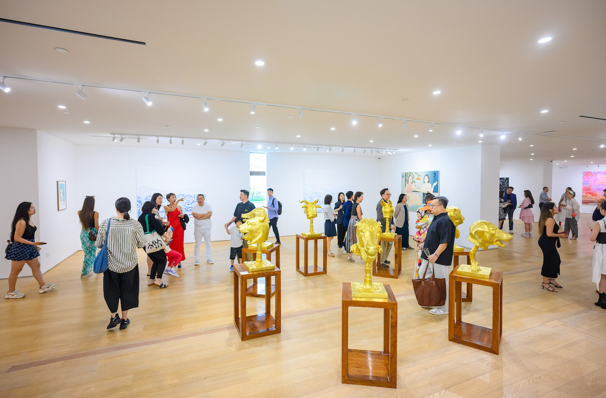 People walk around a gallery with several gold scuptures on wooden plinths in the foreground.