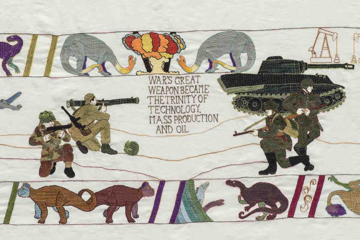 An embroidered textile work with images of soldiers, tanks, and animals and text that reads "war's great weapon became the trinity of technology, mass production and oil."