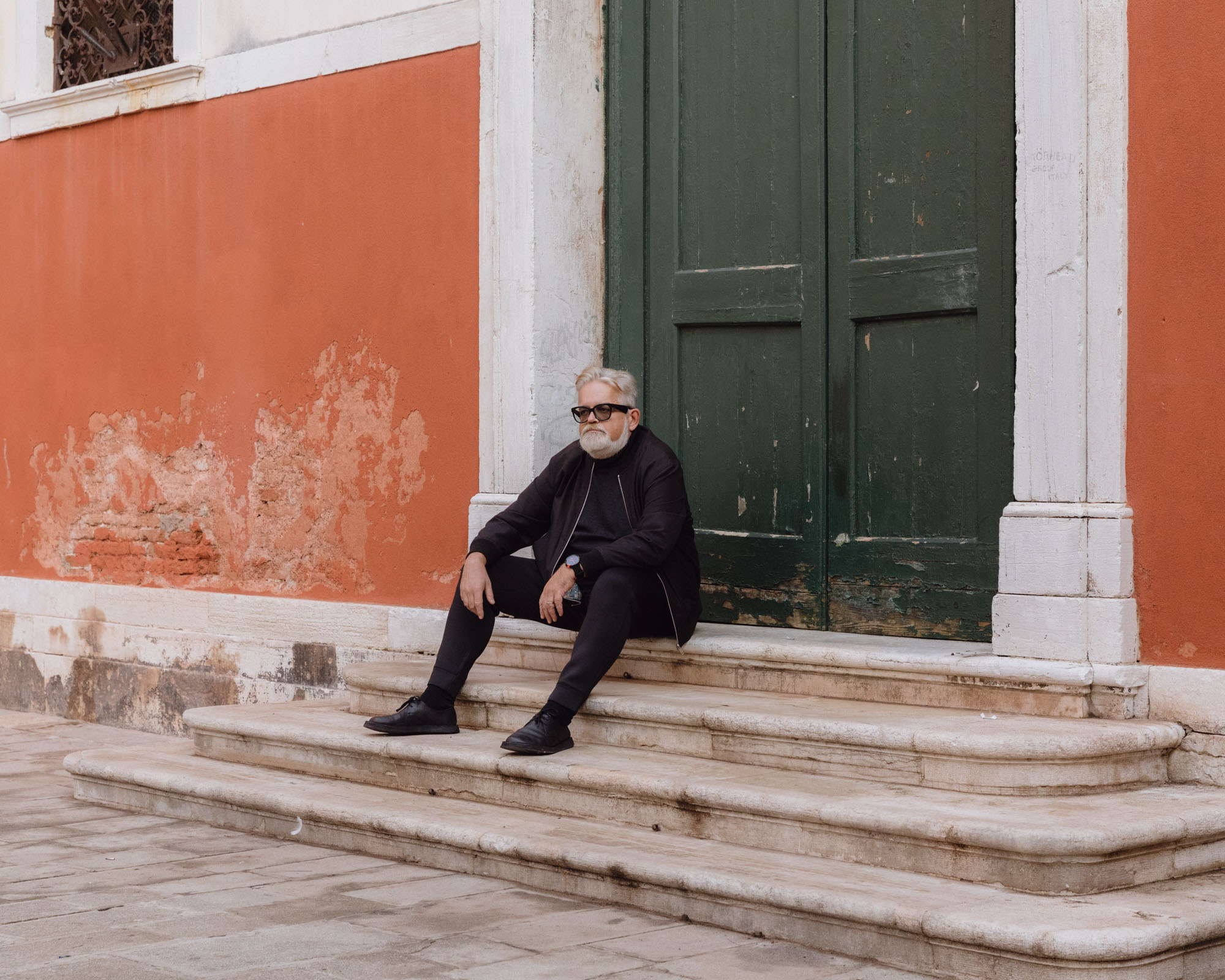 A man dressed in black sits on steps by a large, double door.