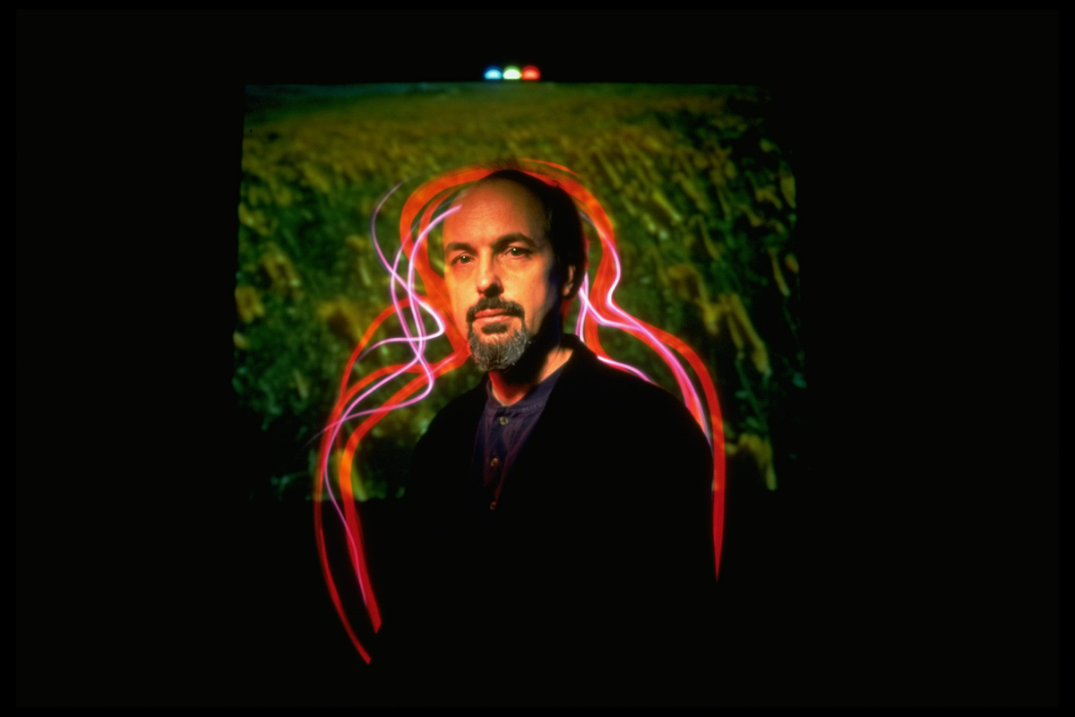 A white man surrounded by red lasers. Behind him is a screen showing a wheat field.