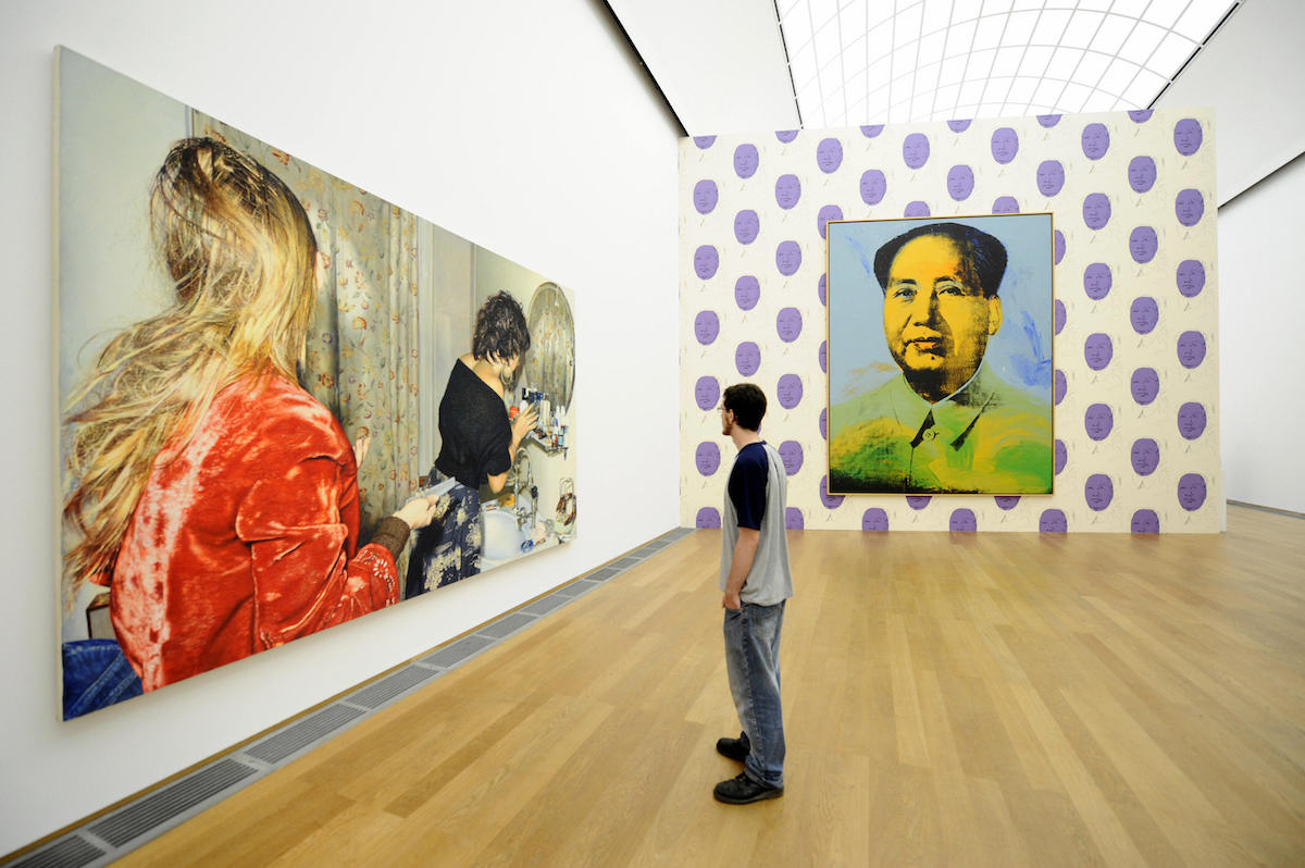 A man staring at a long painting of two women. On an adjacent wall, an image of Chairman Mao hangs above purple polka dots.