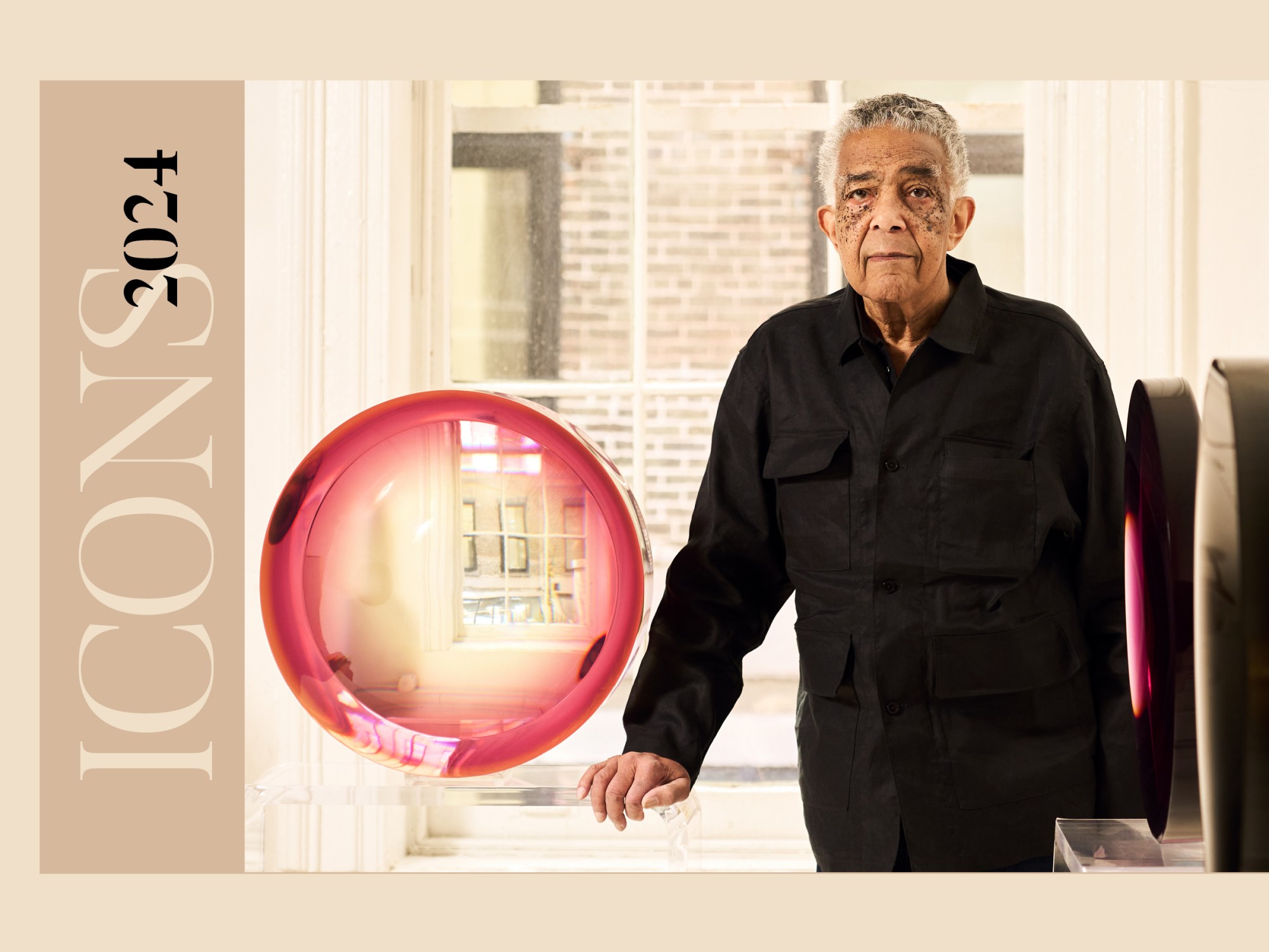 An elderly black man stands next to a clear round sculpture with a pink halo effect. He is wearing all black and resting a hand on a clear pedestal.