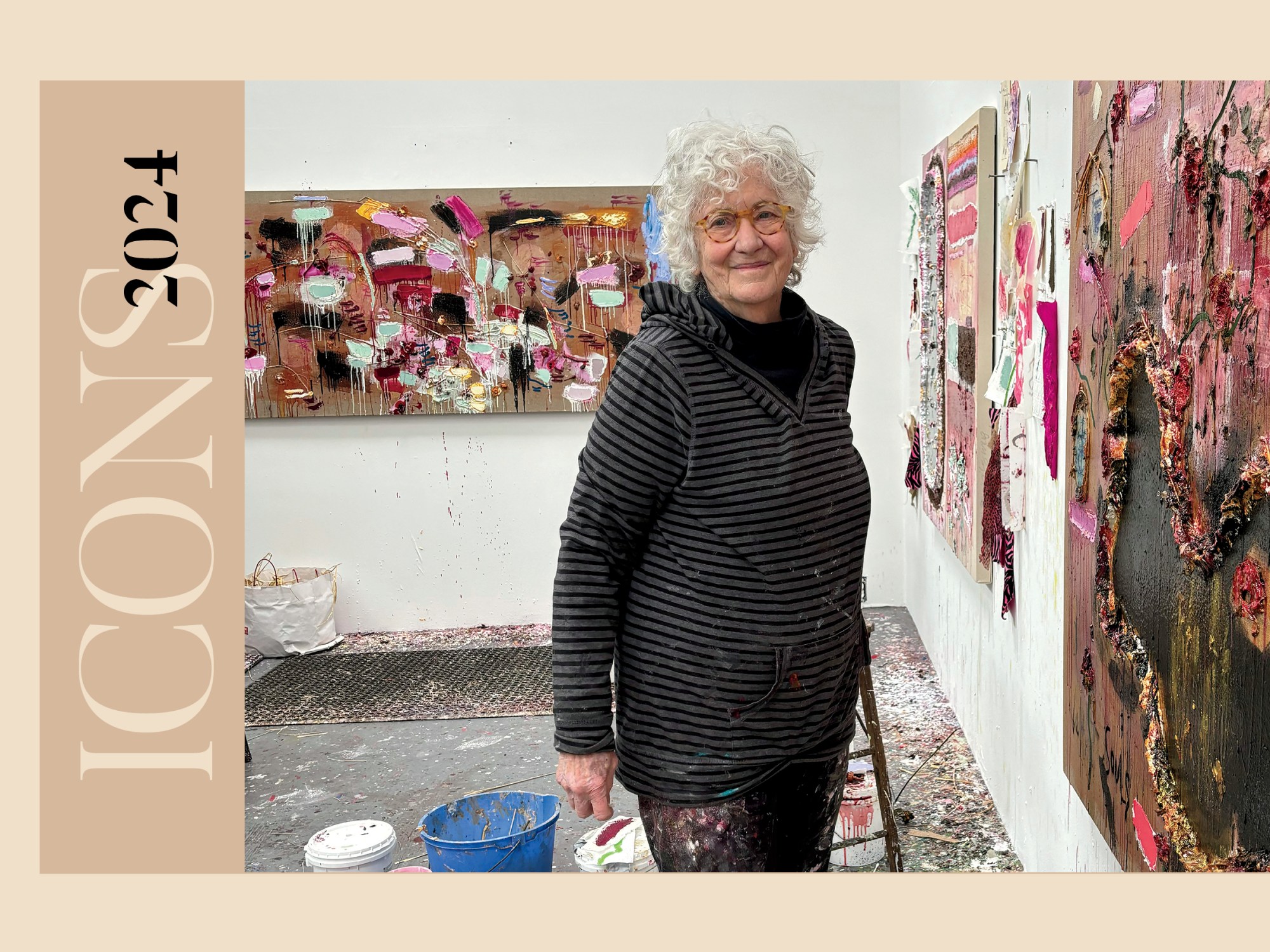 A white woman in a black hooded sweatshirt and black pants surrounded by multi-colored abstract paintings.