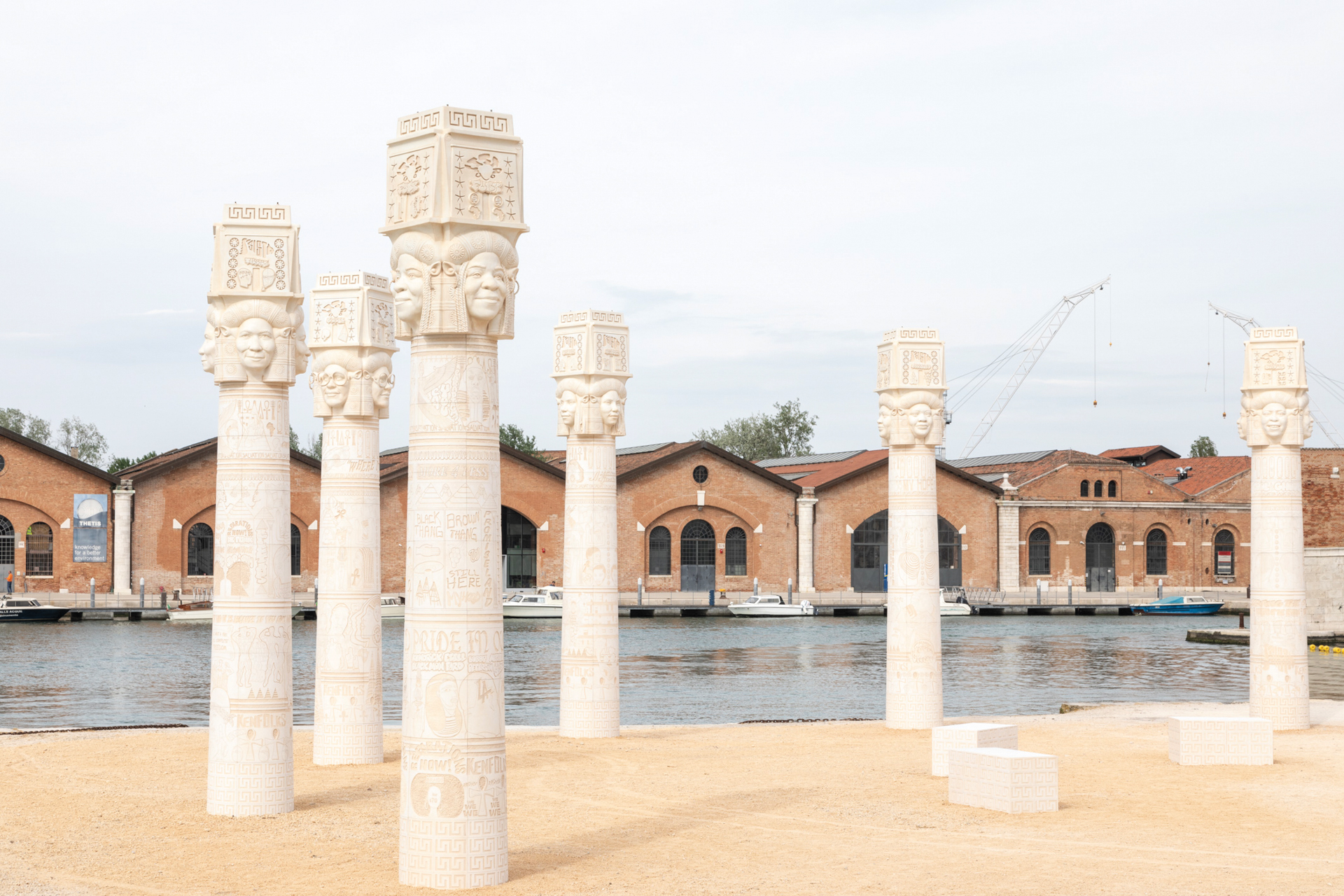 Six egyptian stype columns tower near water. They have vernacular signs engraved into their bases and portraits for capitals.